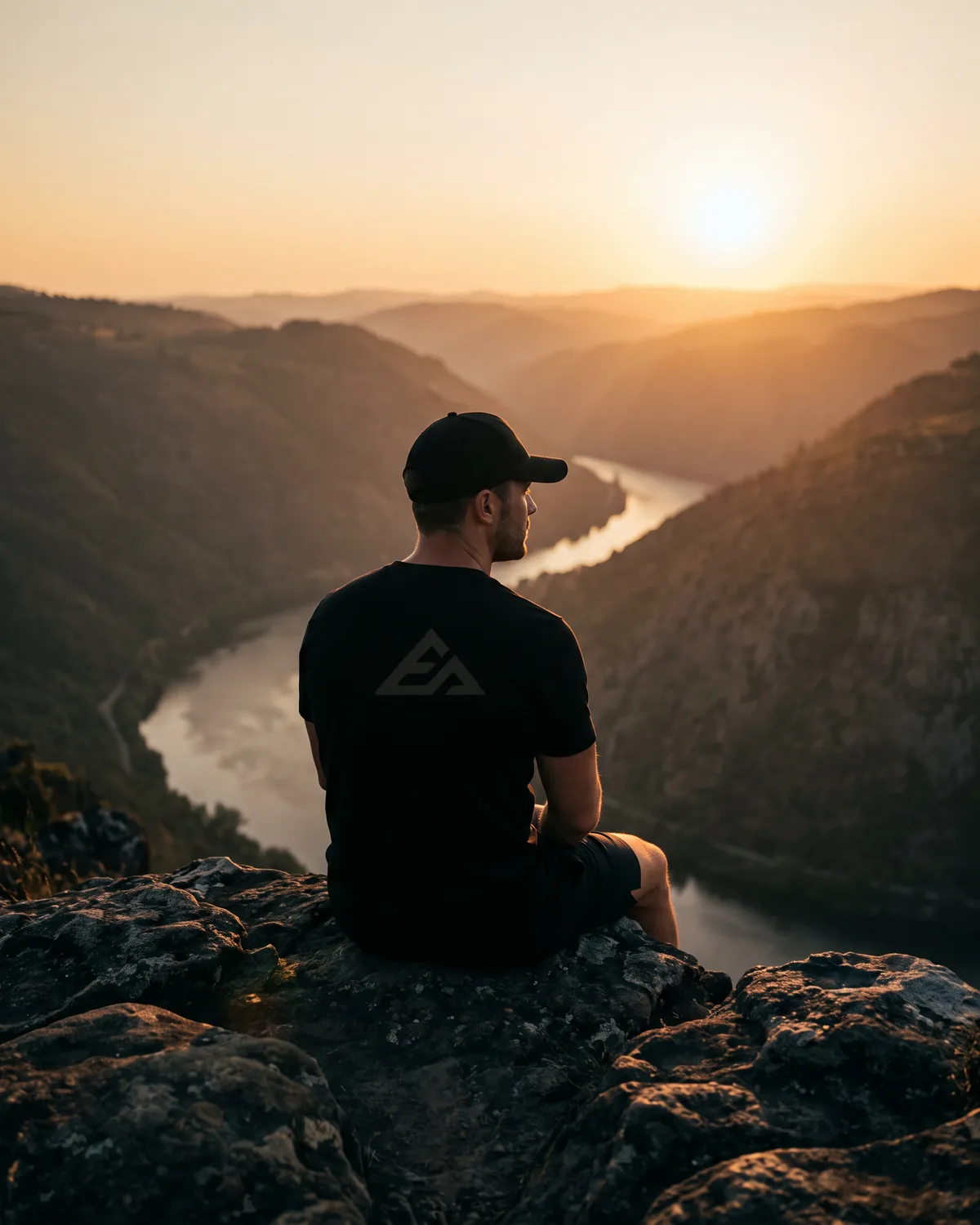 Man in a black tee and cap sitting on a rocky ledge above a river valley at sunset